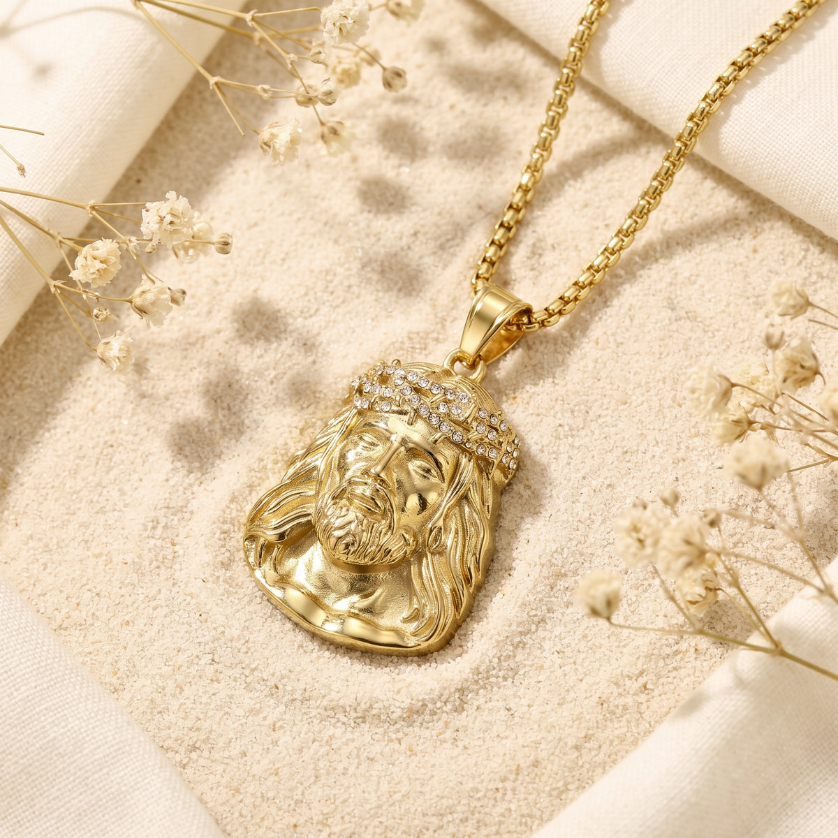Gold-finish Jesus portrait pendant with cubic zirconia crown of thorns resting on sand with dried flowers.
