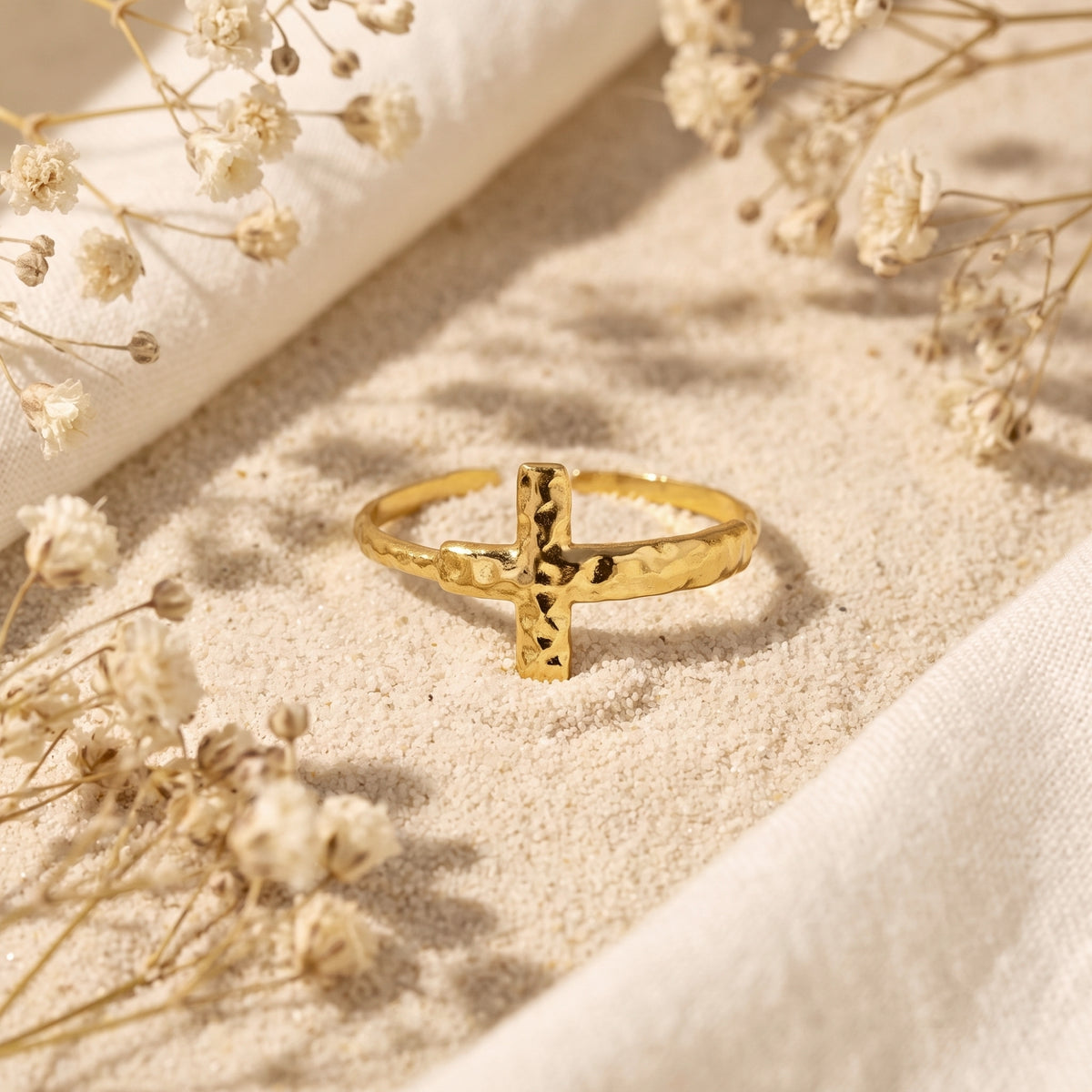 Gold-tone hammered cross ring standing in fine sand among white dried flowers.