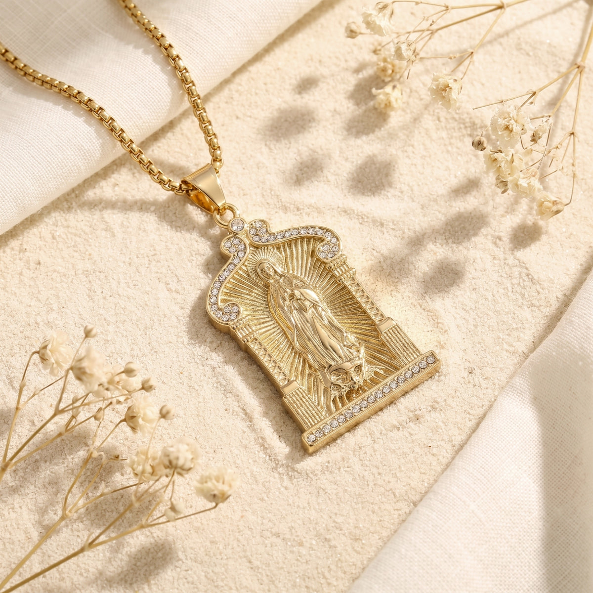 Gold-tone Guadalupe arch pendant with clear crystals lying diagonally on sand beside dried flowers and linen.