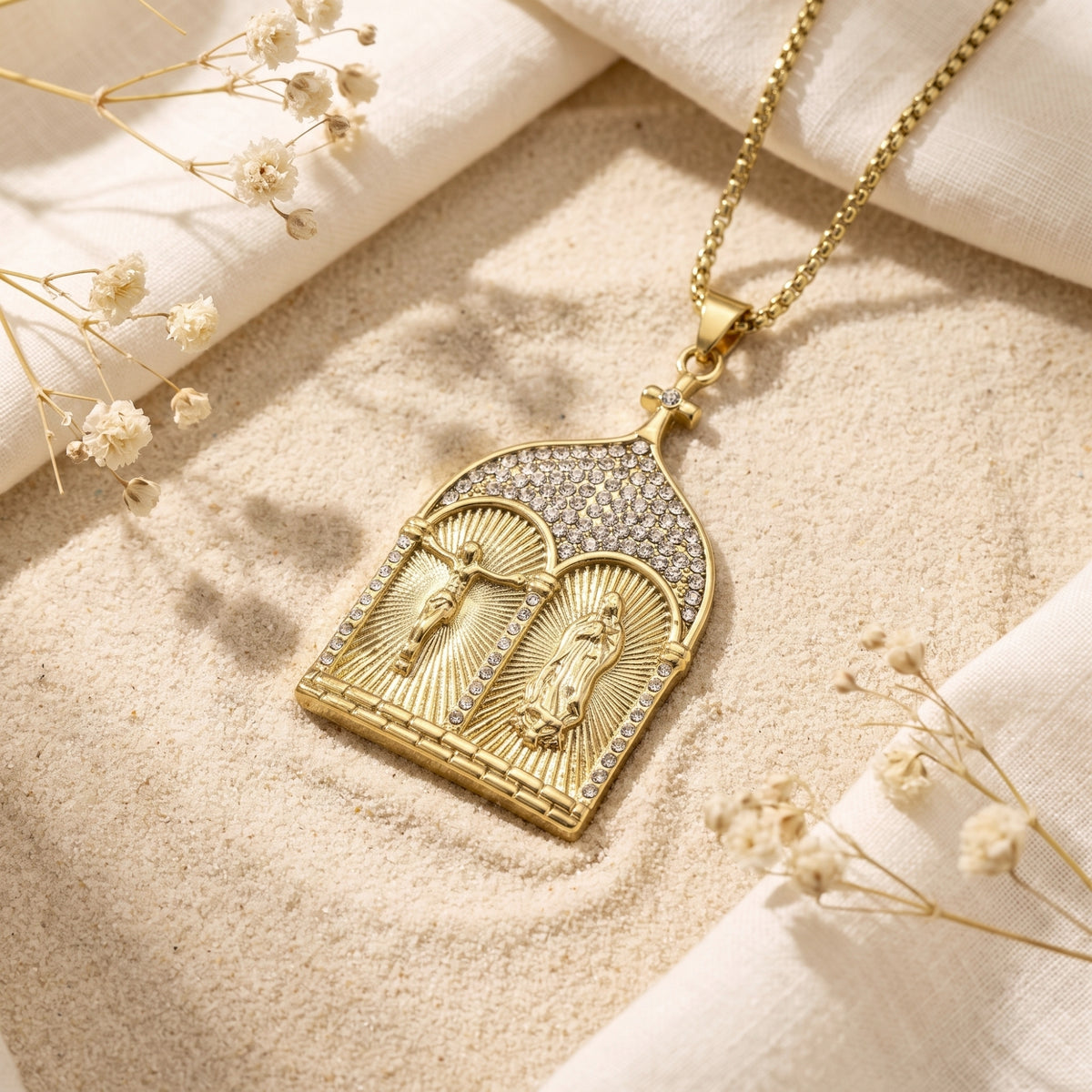 Gold-tone diptych pendant necklace resting on sand with white cloth and dried flowers.