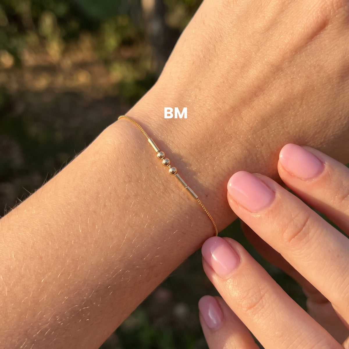 Gold-finish Morse-code bracelet worn on a wrist, with the BM example label.
