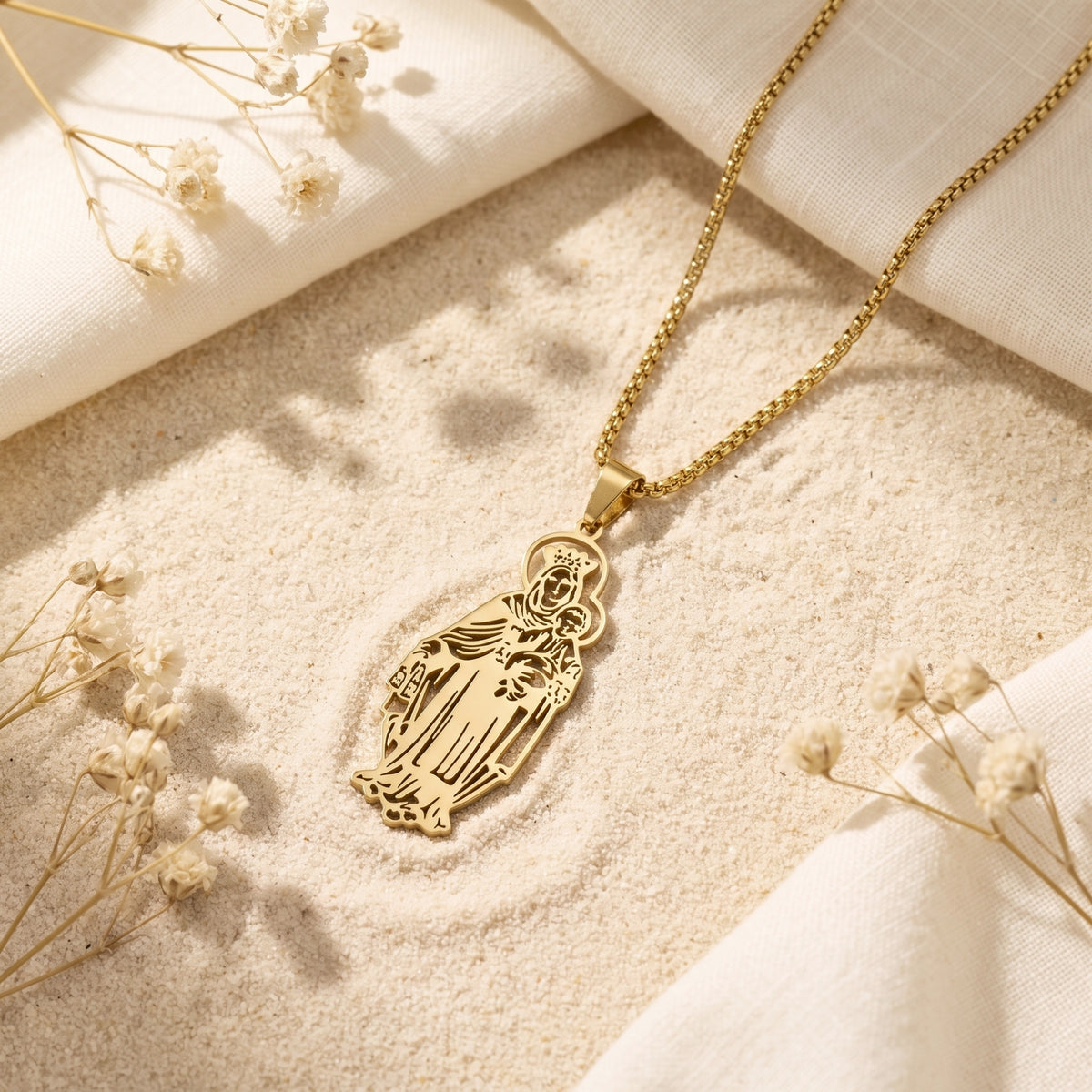 Gold Madonna and Child pendant necklace resting on sand beside white fabric and dried flowers.