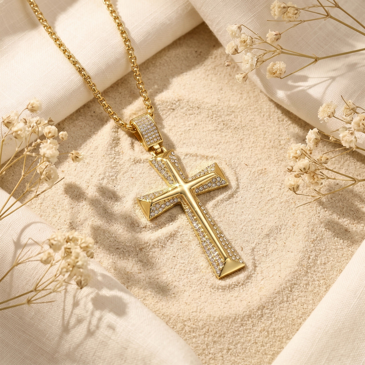 Gold-tone beveled cross pendant with pavé accents and chain resting diagonally on sand among white fabric and dried flowers.