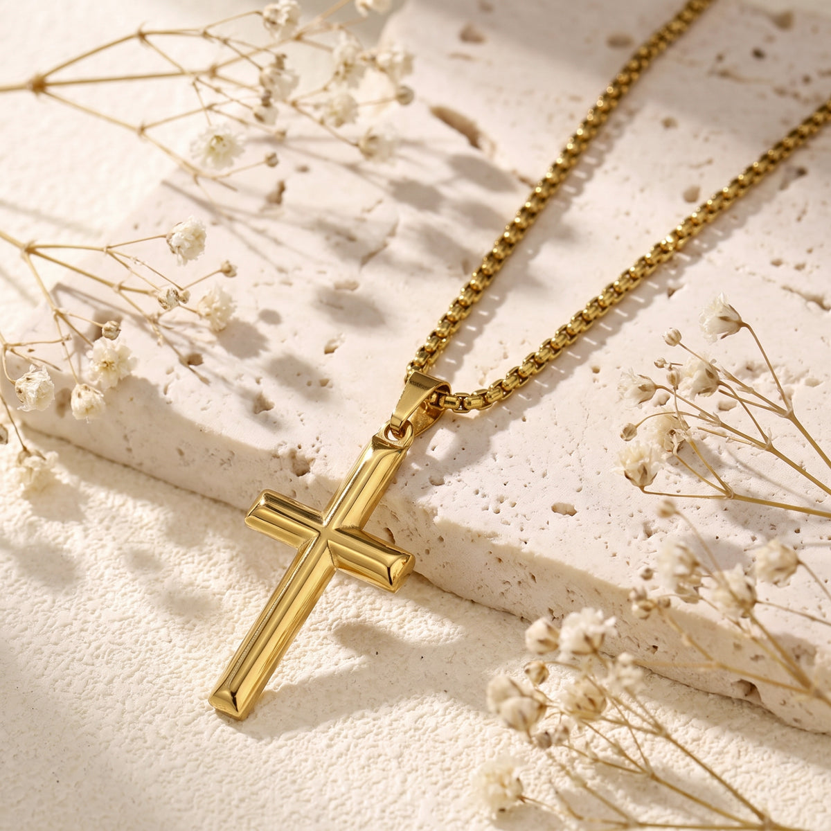 Gold-tone architectural cross pendant and chain lying on a textured stone surface near dried flowers.