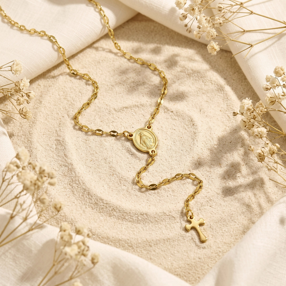 Flat lay of the gold-tone rosary cross necklace with Miraculous Medal and cross on sand with dried flowers.