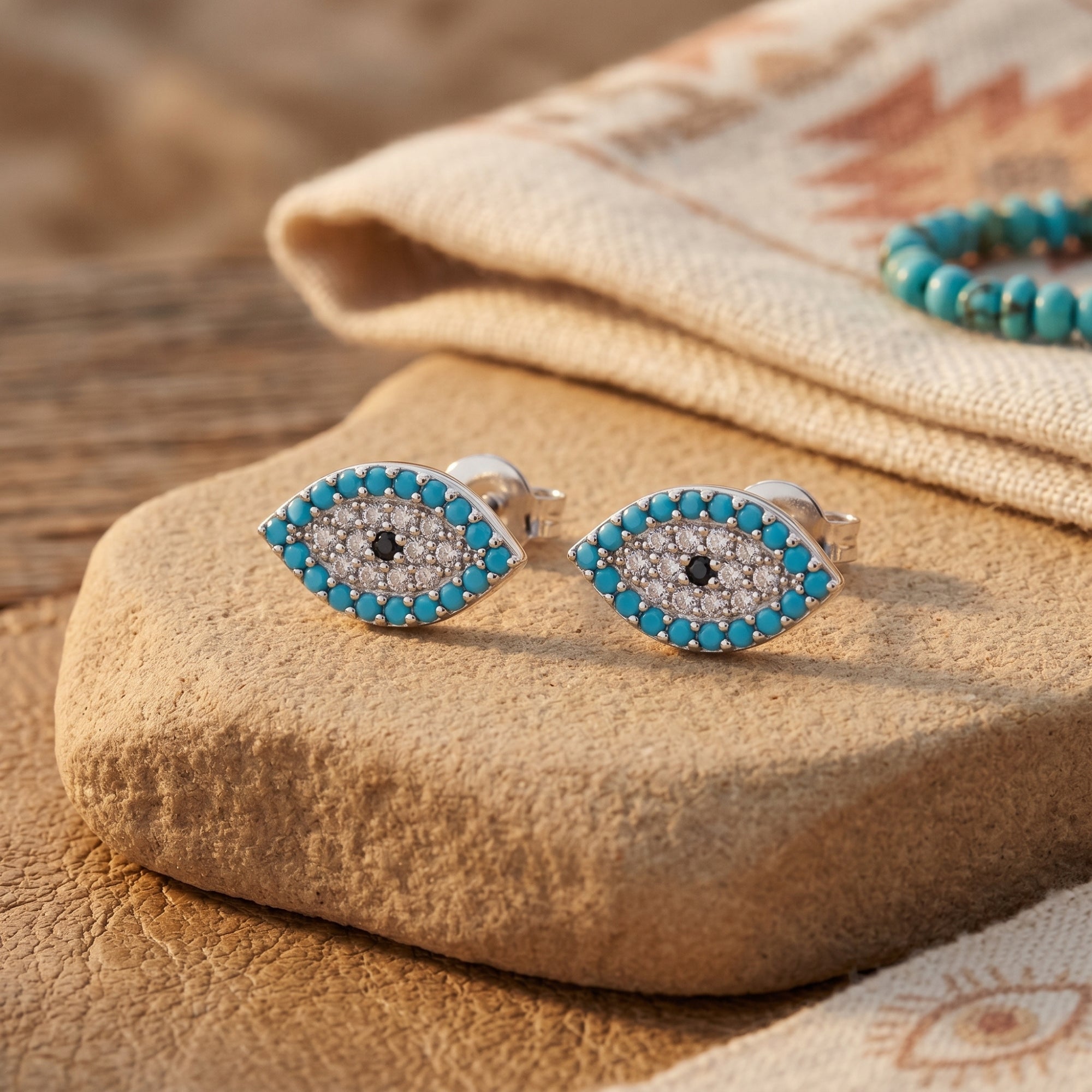 Silver turquoise evil eye stud earrings with simulated diamonds resting on a stone in a Southwestern setting.