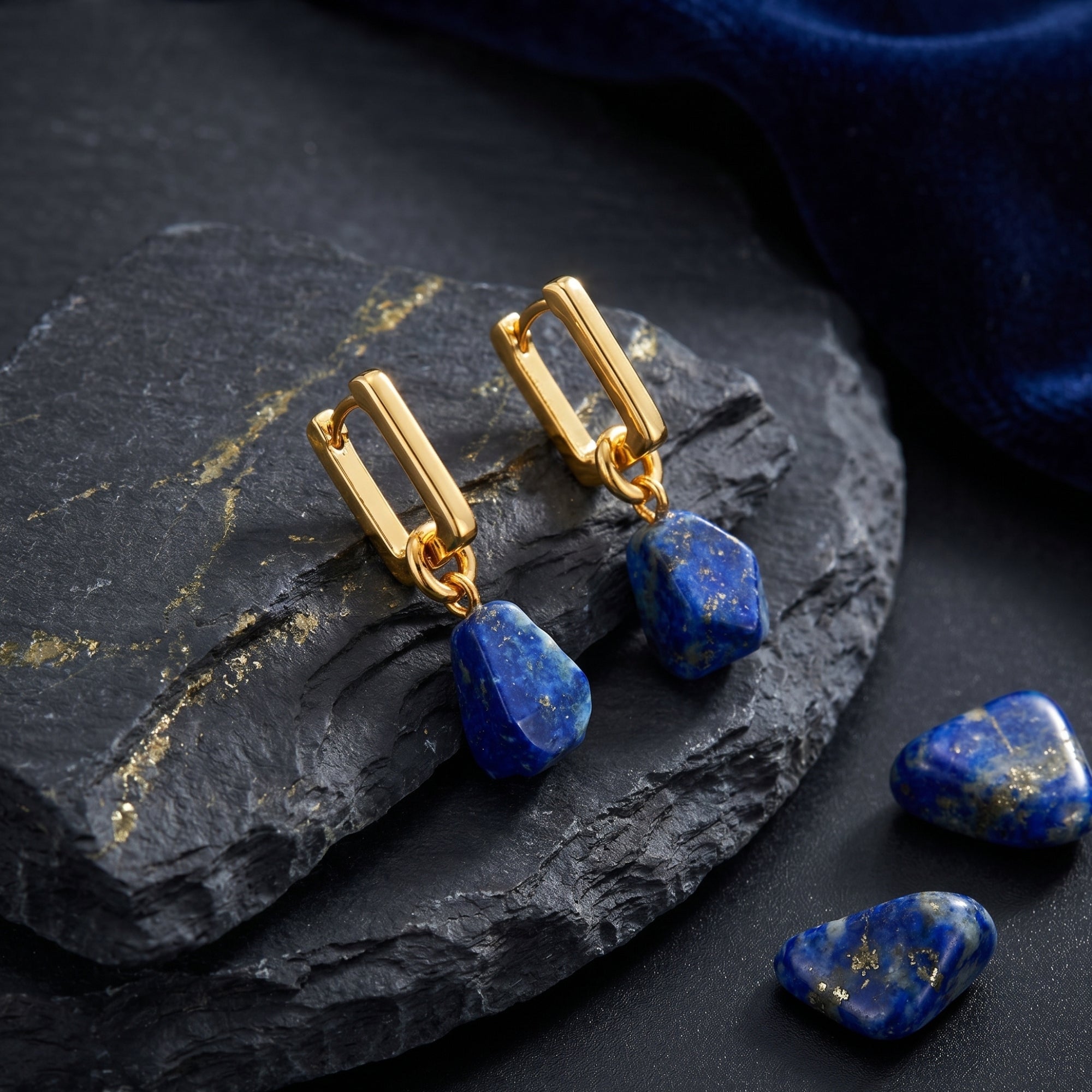 Gold-plated rectangular hoop earrings with irregular lapis lazuli drops styled on a dark rock with blue velvet.