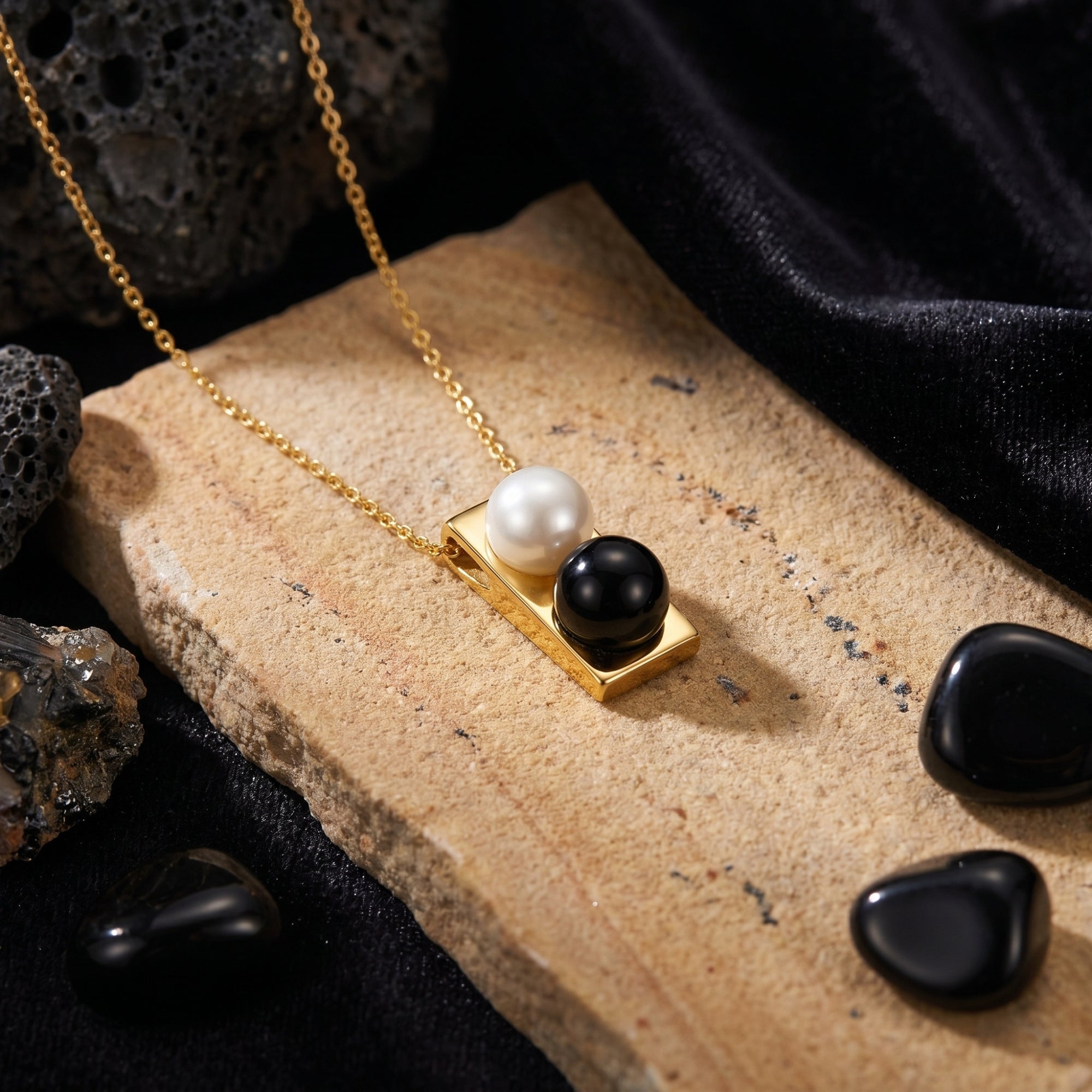 Gold pendant necklace with a white pearl and black onyx cabochon on a textured stone and black velvet background.