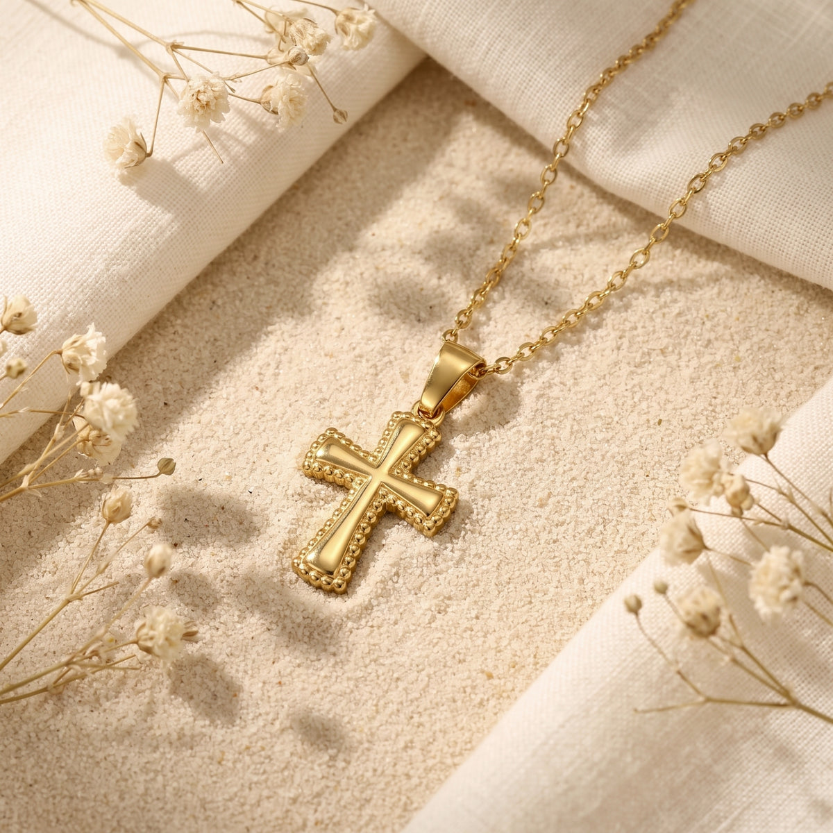 Gold-tone cross pendant necklace with beaded edges resting on fine sand beside white dried flowers.