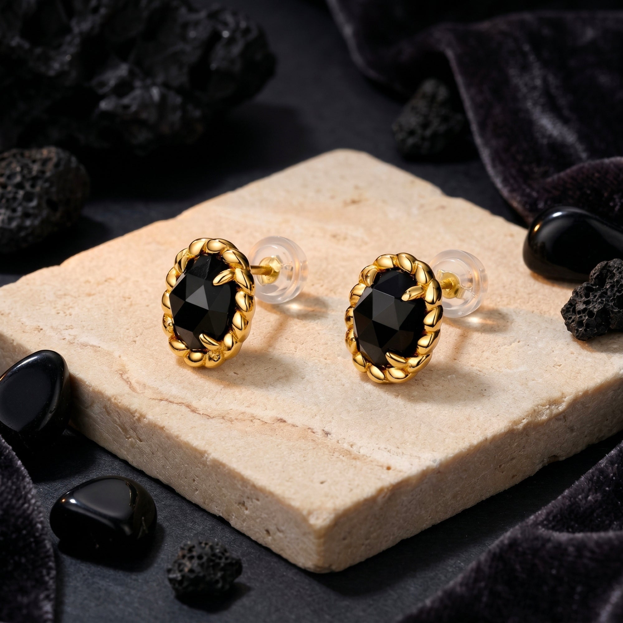 Faceted oval onyx stud earrings in 18K gold plated twist frames styled on a stone slab with black velvet.