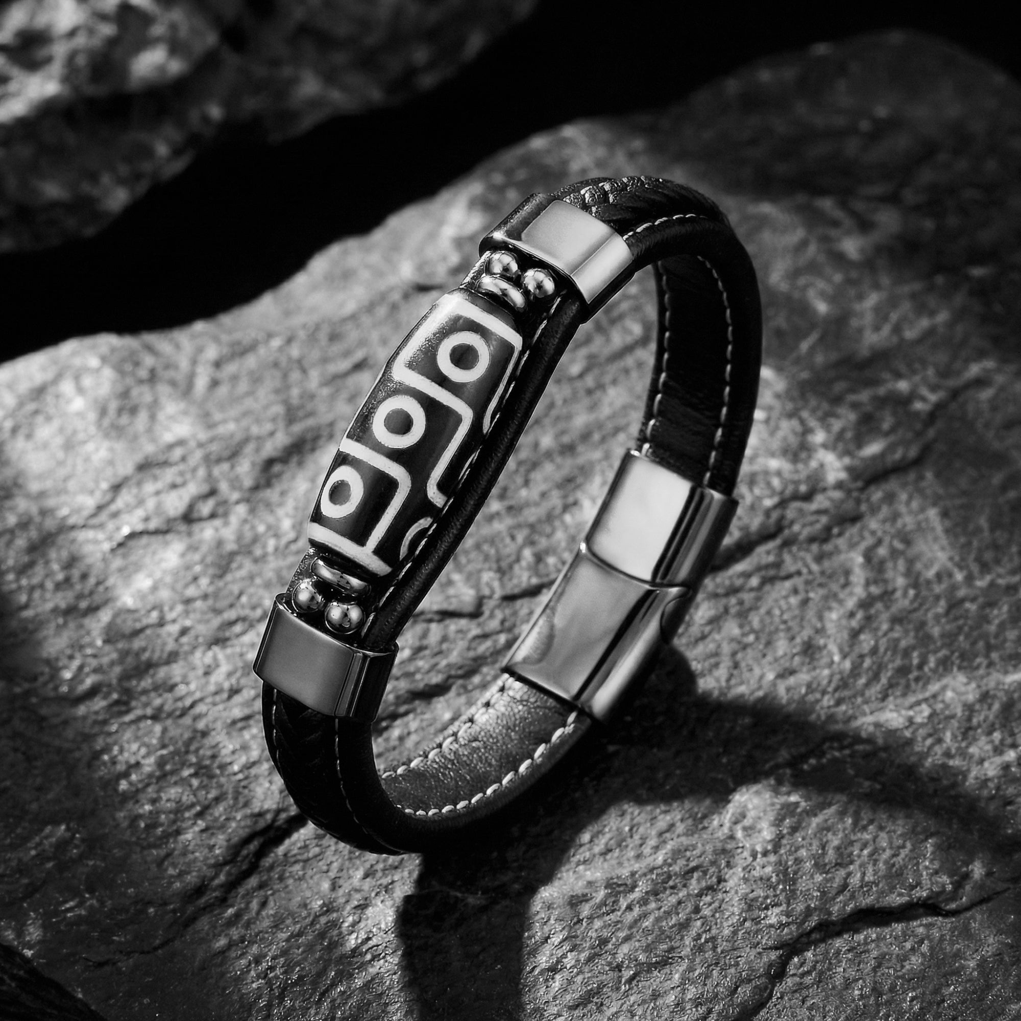 Black braided leather bracelet with a silver-tone 316L stainless steel geometric centerpiece on dark textured stone.