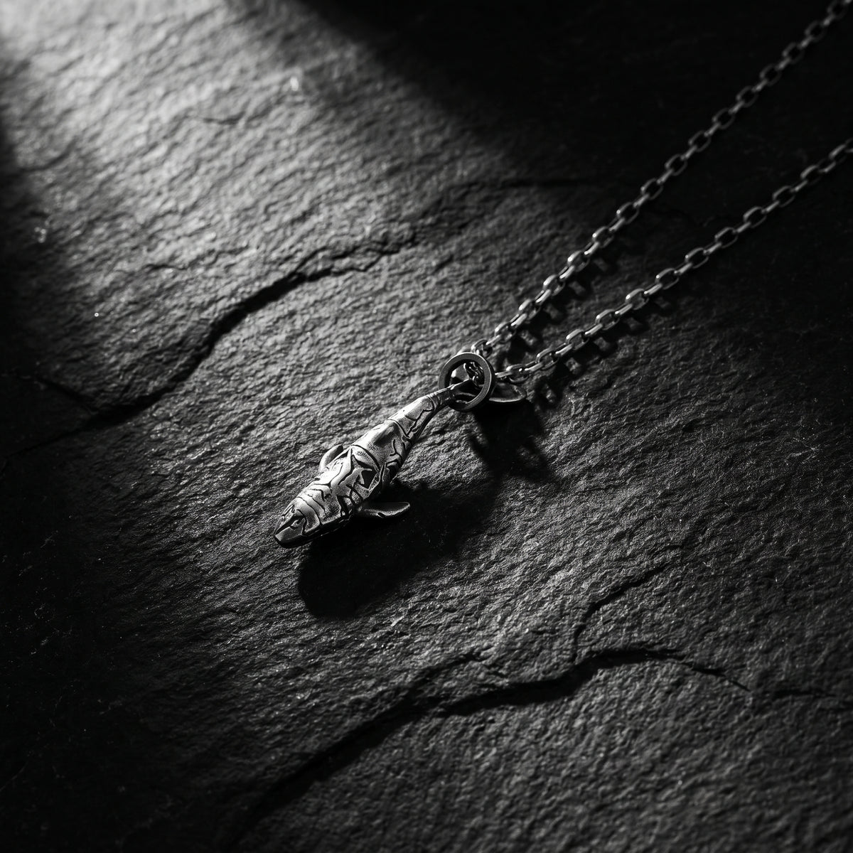 Oxidized silver shark pendant lying diagonally on a dark slate surface with its chain extending outward.