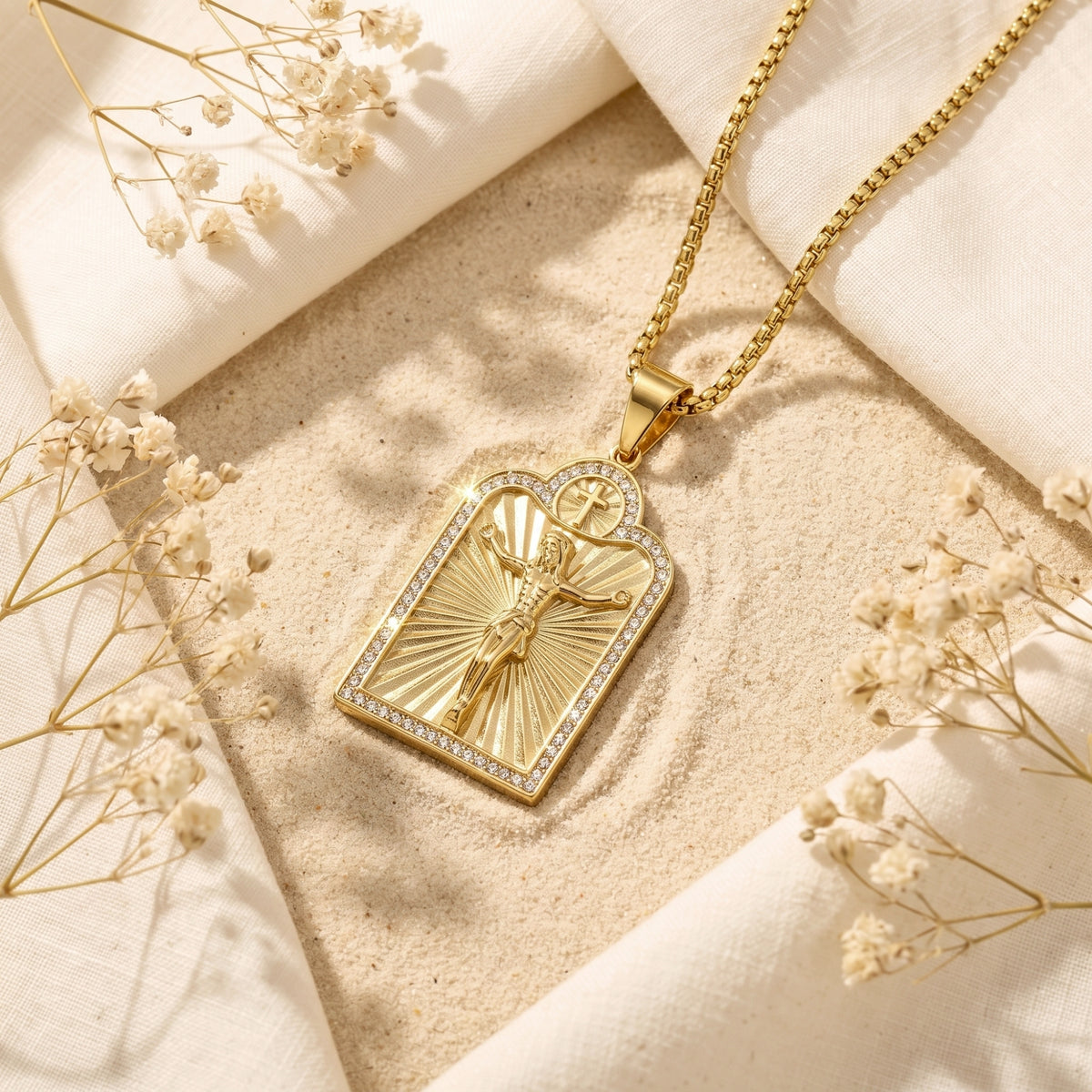 Gold-tone arched crucifix necklace lying on sand, framed by linen fabric and dried flowers.