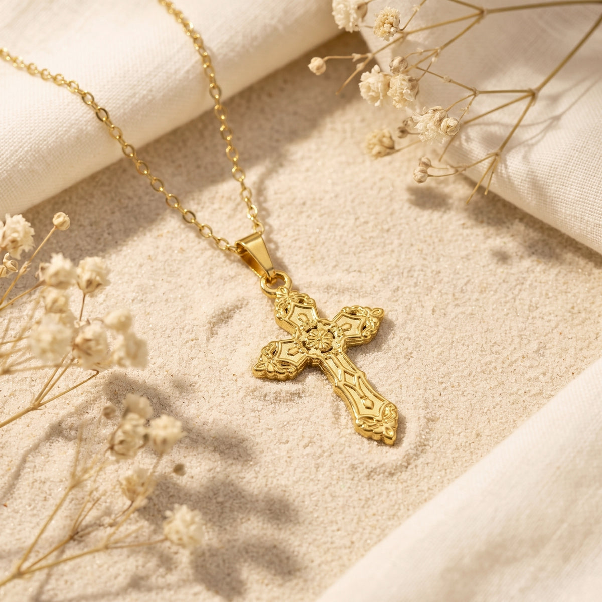 Gold-tone ornate cross pendant and chain resting on sand beside white fabric and dried flowers.