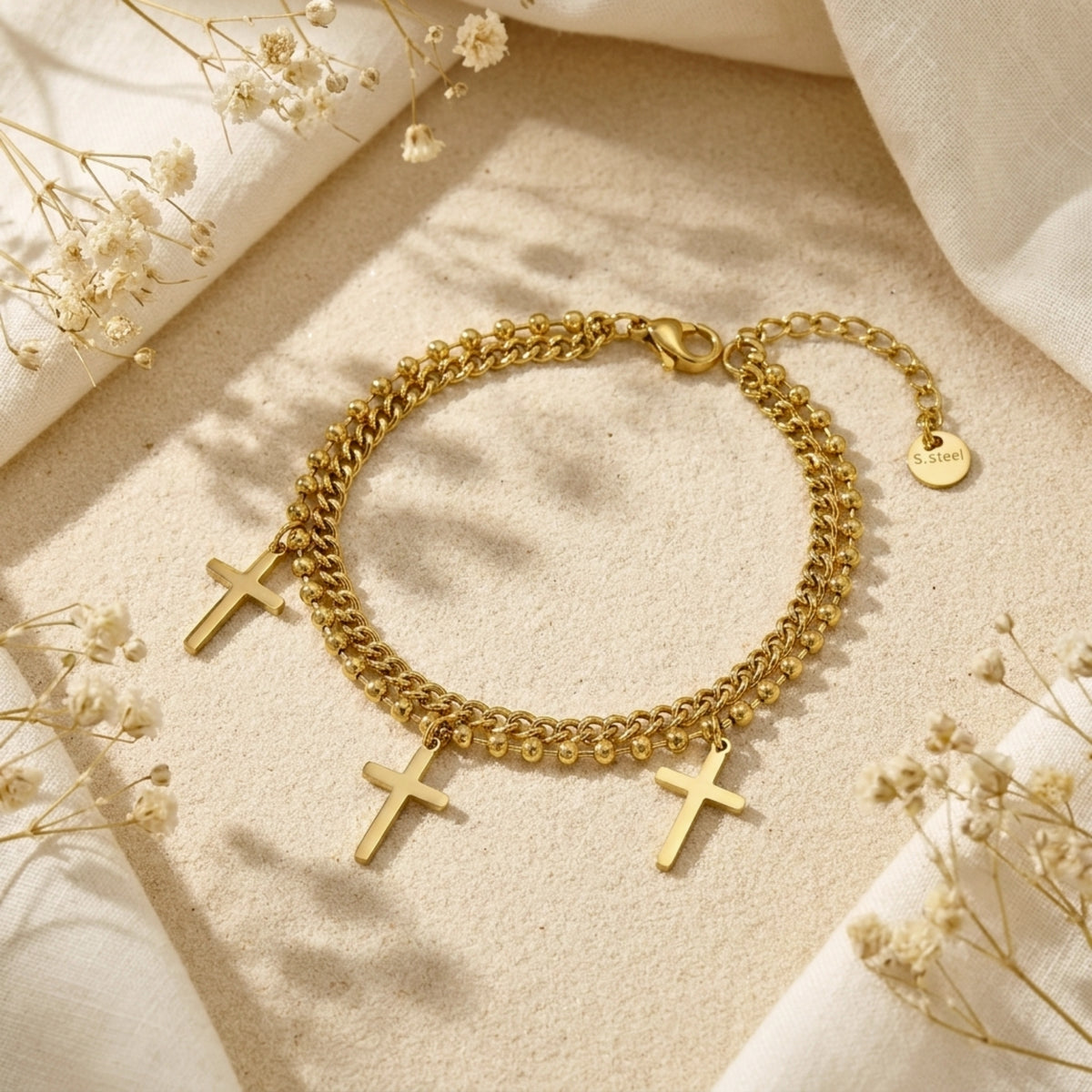 Gold double-strand cross charm bracelet displayed on a sandy surface with white fabric and dried flowers.