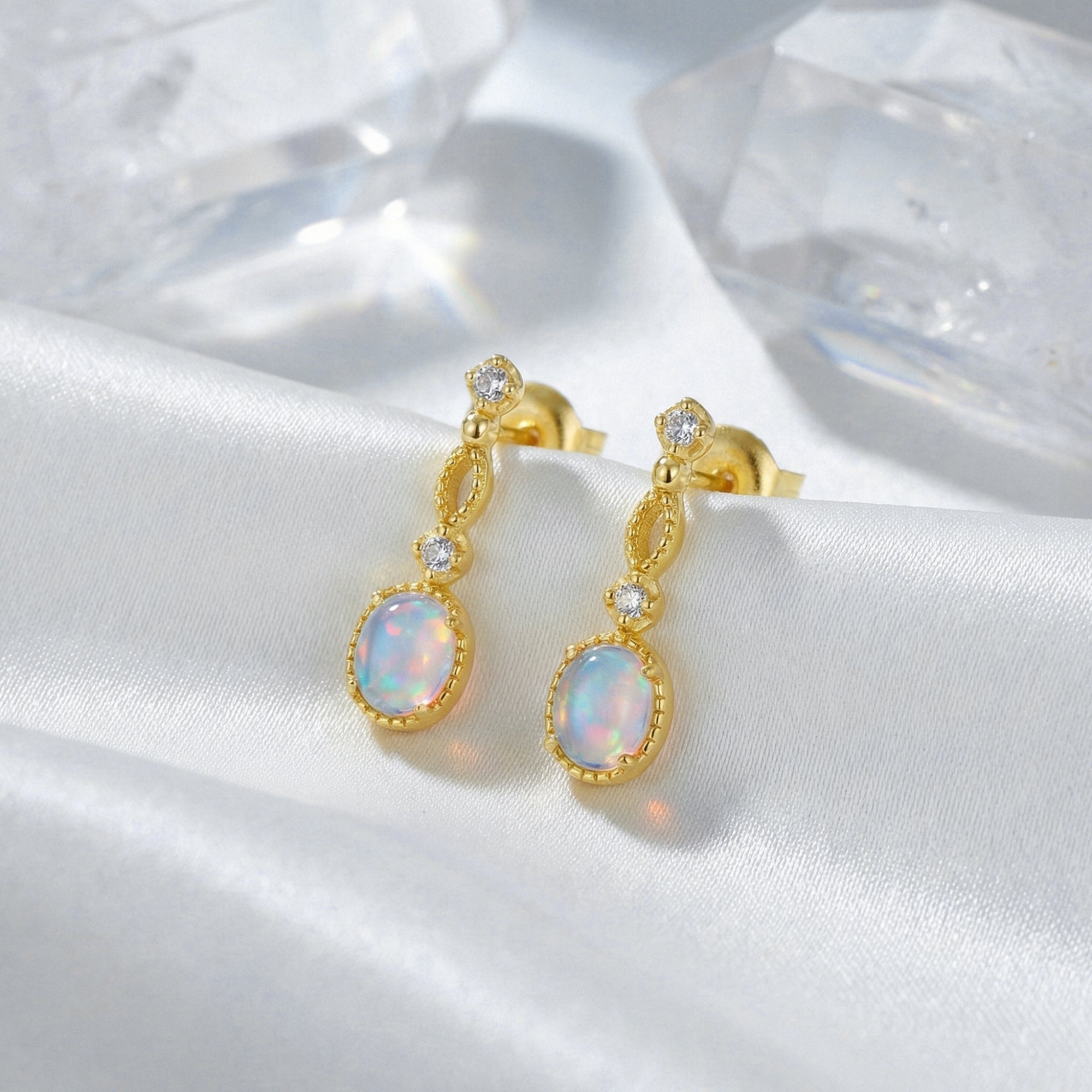 Gold-tone Lorelei drop earrings with oval white opals displayed on white silk with crystal props.