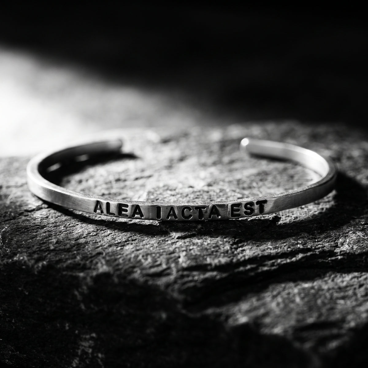 Close-up of a silver open cuff engraved “ALEA IACTA EST” resting on a textured rock surface.