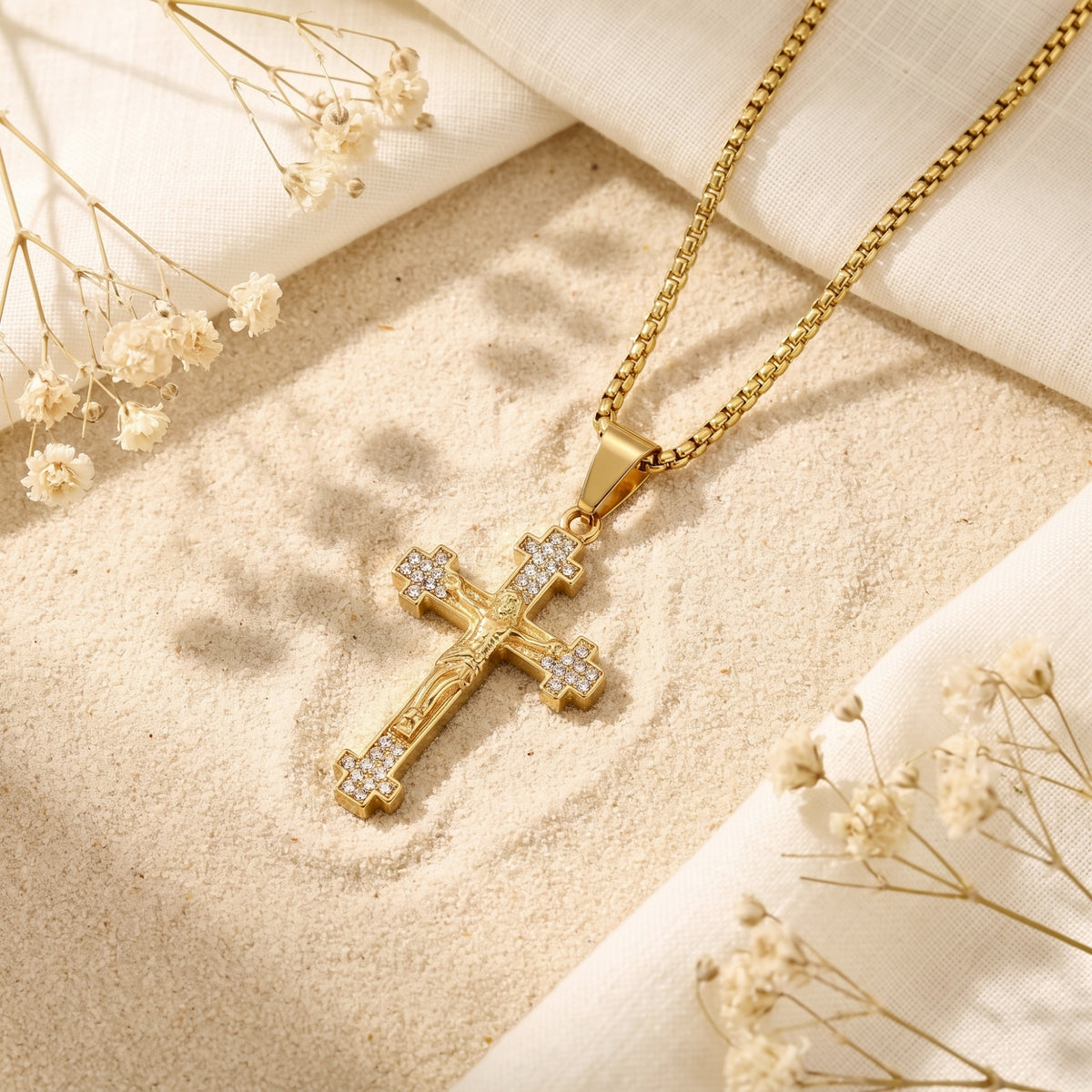 Gold crucifix necklace with clear crystal accents lying on sand among white linen and dried flowers.