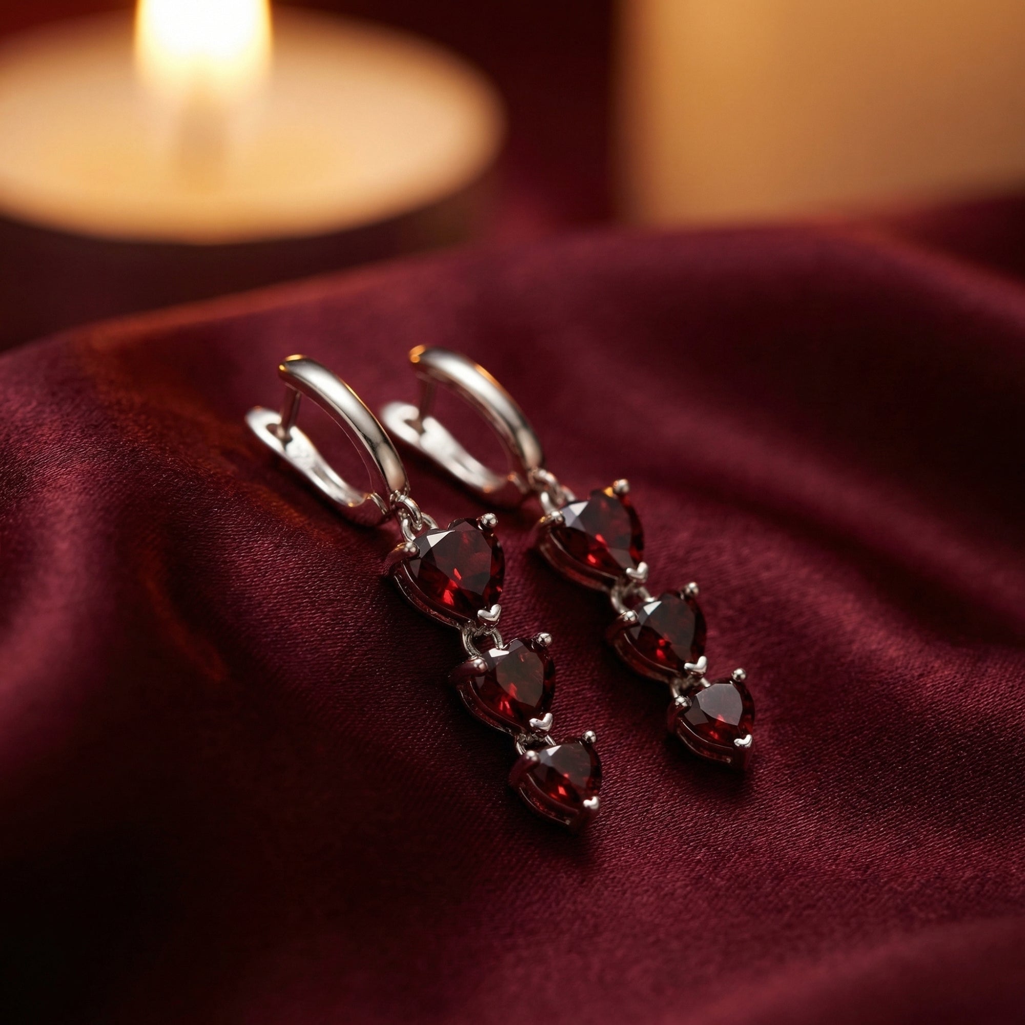 Silver heart drop earrings with three tiered heart-cut red garnets on burgundy silk with a candle background.