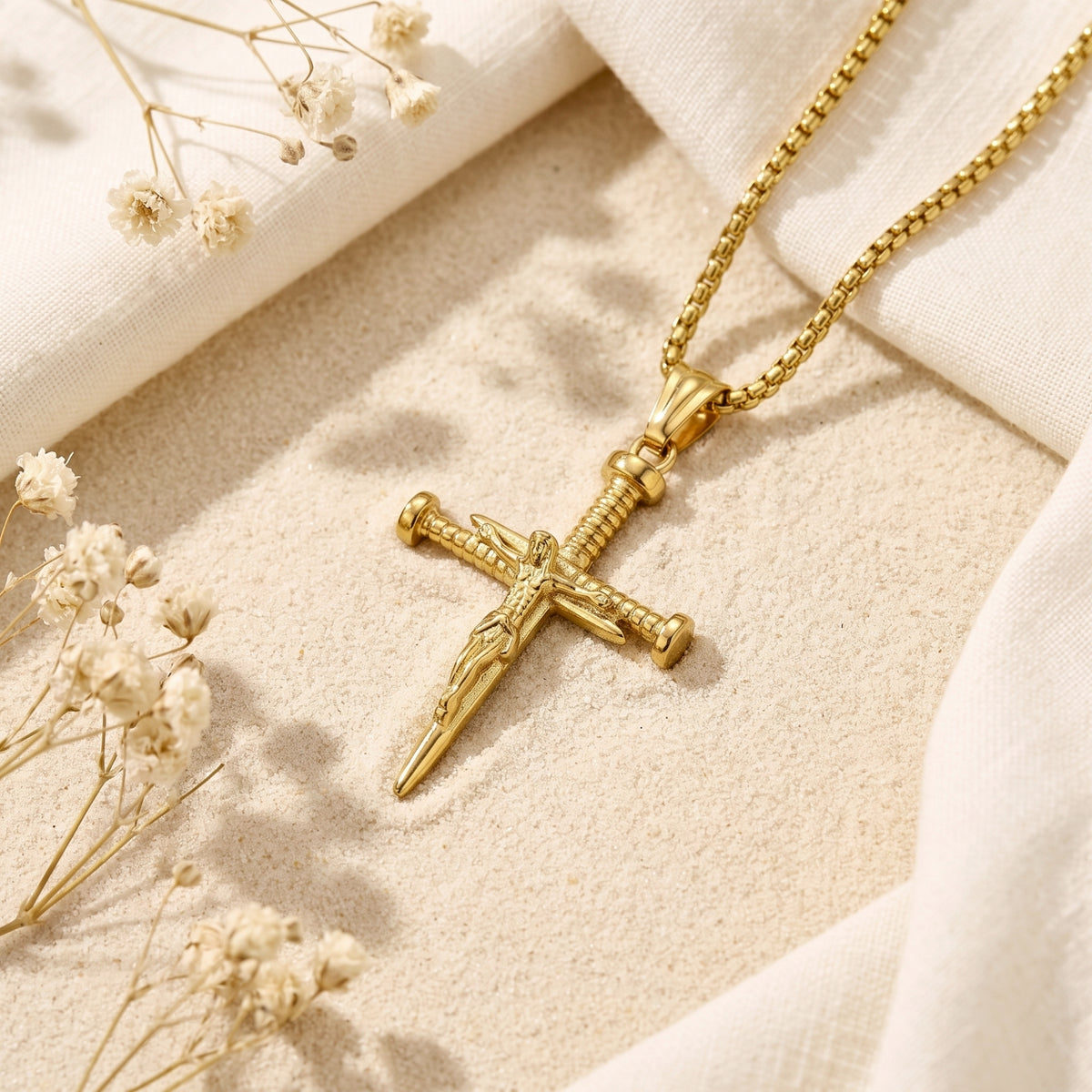 Gold-tone nail crucifix pendant with a box chain resting on sand among white dried flowers and fabric.