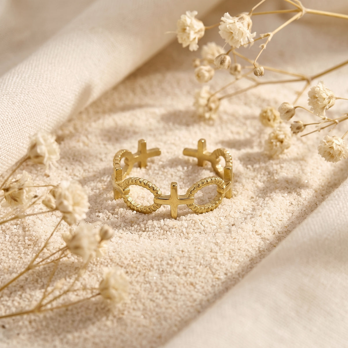 Gold-tone open cross ring with rope-textured bands resting on white sand among dried flowers.