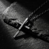 Silver textured cross pendant and chain lying diagonally on a dark slate surface.