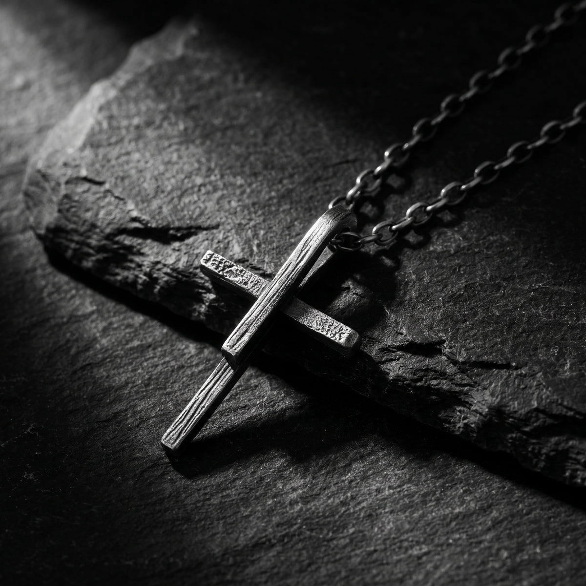 Silver textured cross pendant and chain lying diagonally on a dark slate surface.