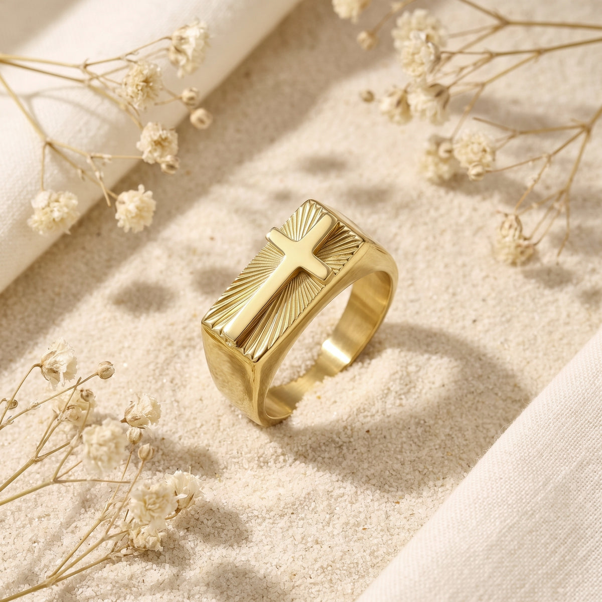 Gold-tone signet ring with a raised cross and sunburst face standing on white sand among dried flowers.