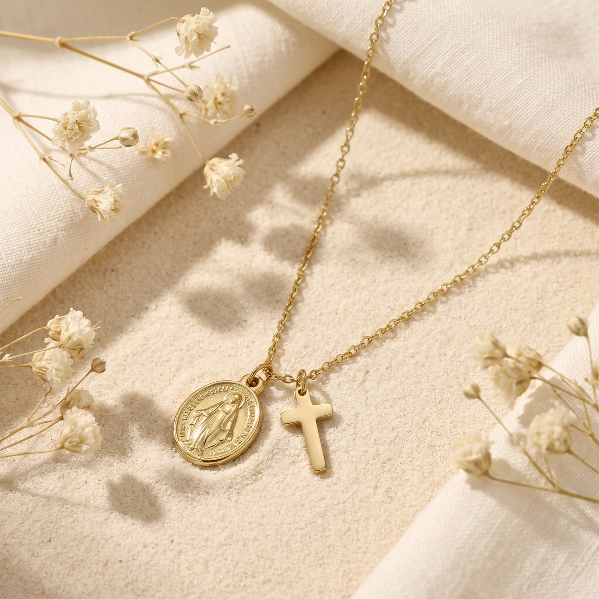 Gold-tone cross and oval medal necklace lying flat on sand with white fabric and dried flowers.