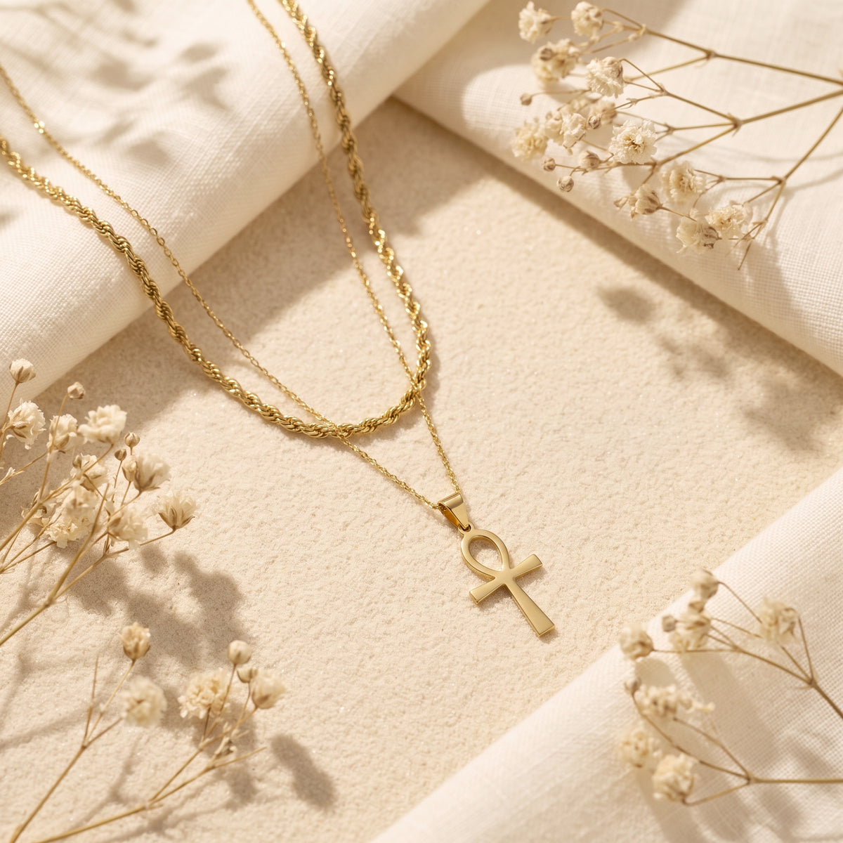 Layered gold-tone Ankh necklace with rope and thin chains laid flat on sand with linen fabric and dried flowers.