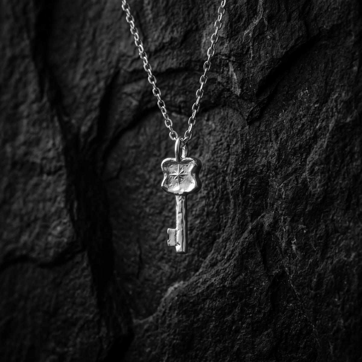 Silver key pendant with a starburst emblem hanging vertically on a chain against dark textured rock.