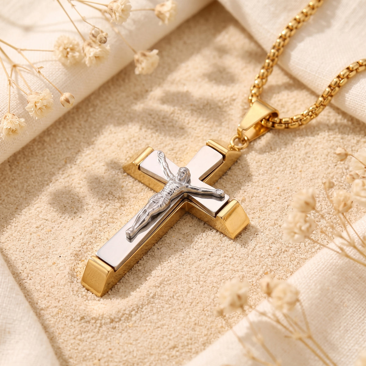Two-tone layered crucifix pendant lying diagonally on sand, with its gold-tone chain and dried flowers nearby.