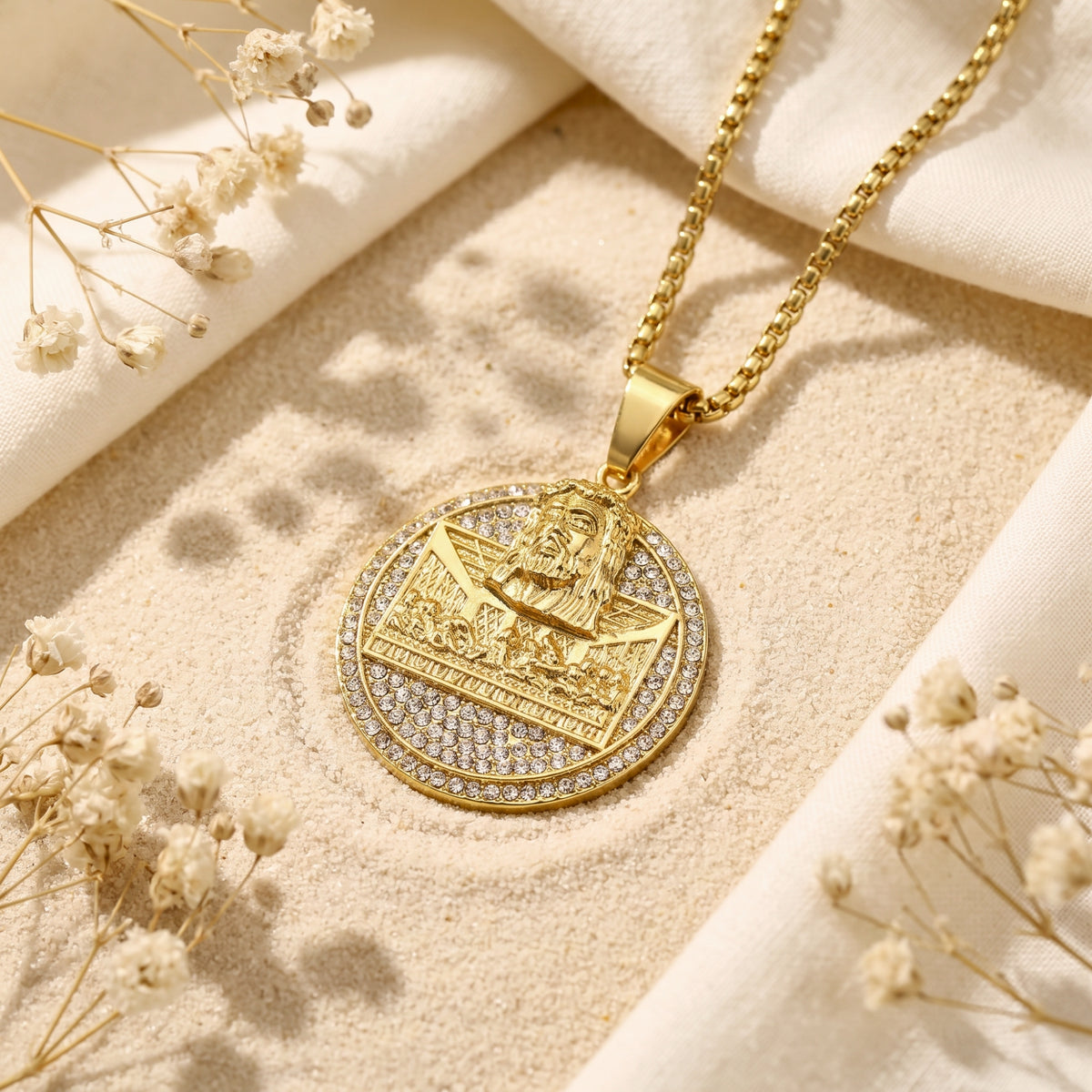 Gold-tone round Last Supper medallion pendant with crystal accents resting on sand among white cloth and dried flowers.