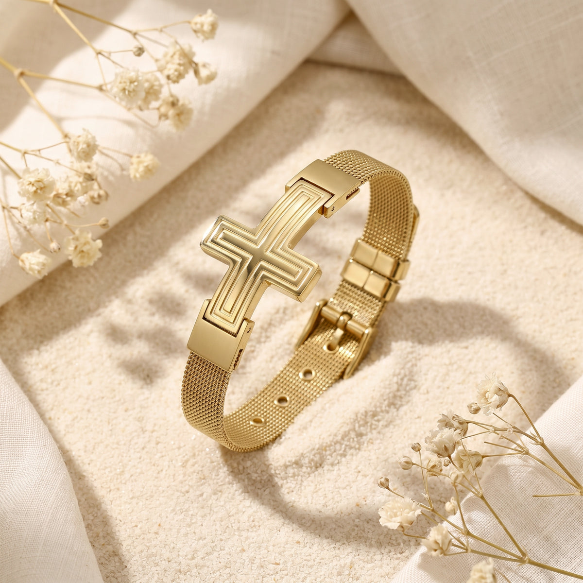 Gold-tone mesh bracelet with an etched cross motif lying at an angle on sand with dried flowers.