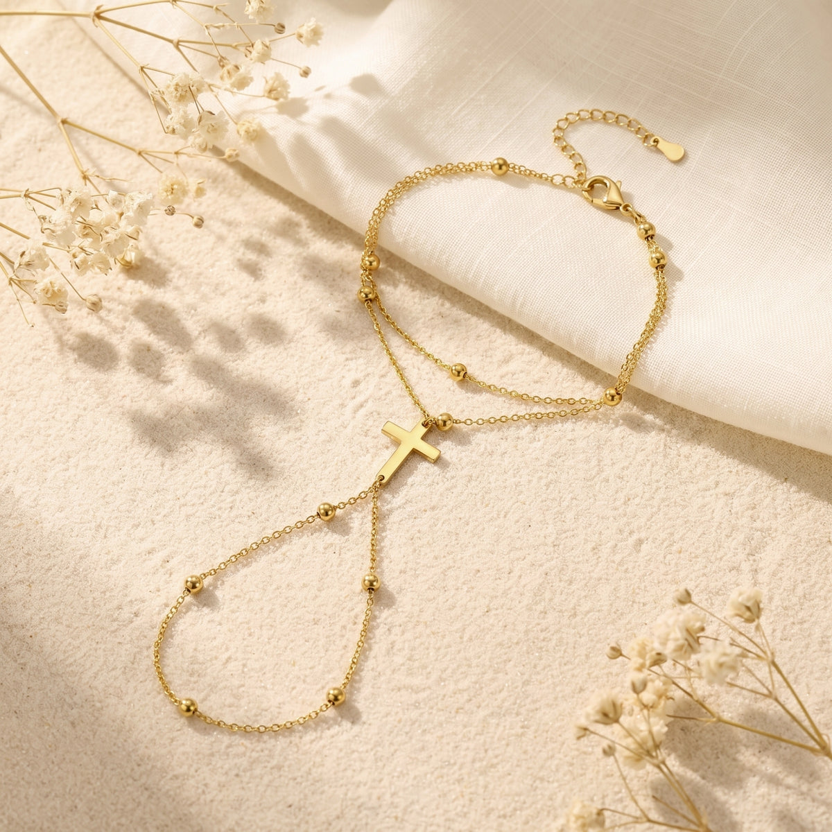 Gold cross hand chain bracelet with beaded stations laid flat on sand beside white fabric and dried flowers.