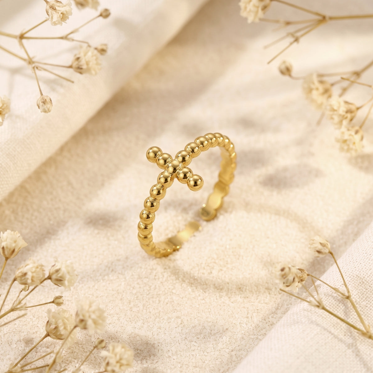 Angled close-up of a gold-toned beaded cross ring resting on fine sand with dried white flowers nearby.