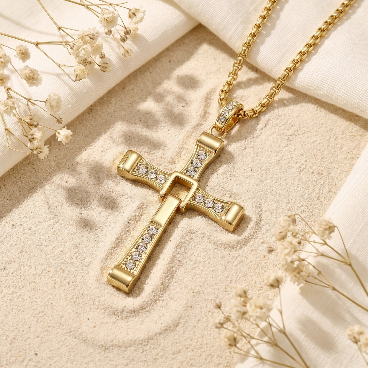 Gold-tone hinged cross pendant with clear crystals lying on sand beside dried flowers and white cloth.