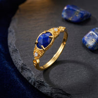 Gold Elarissa ring with a faceted oval lapis lazuli stone styled on a dark stone slab with loose blue gems.
