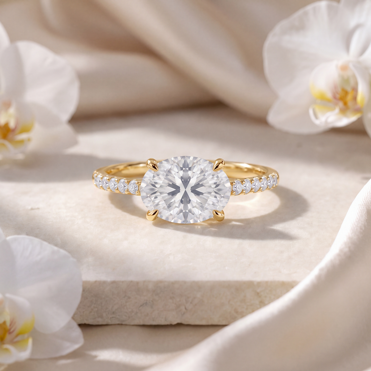 Yellow gold east-west oval moissanite engagement ring with a pave diamond band resting on a stone surface.