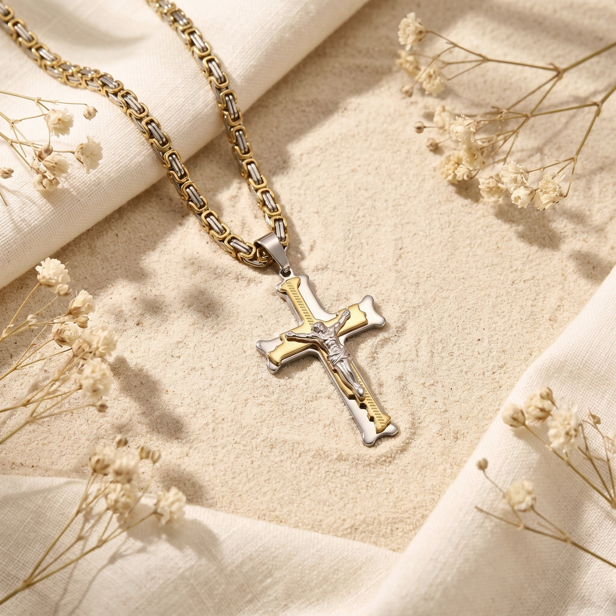 Two-tone crucifix pendant and chain resting flat on sand alongside white fabric and dried flowers.