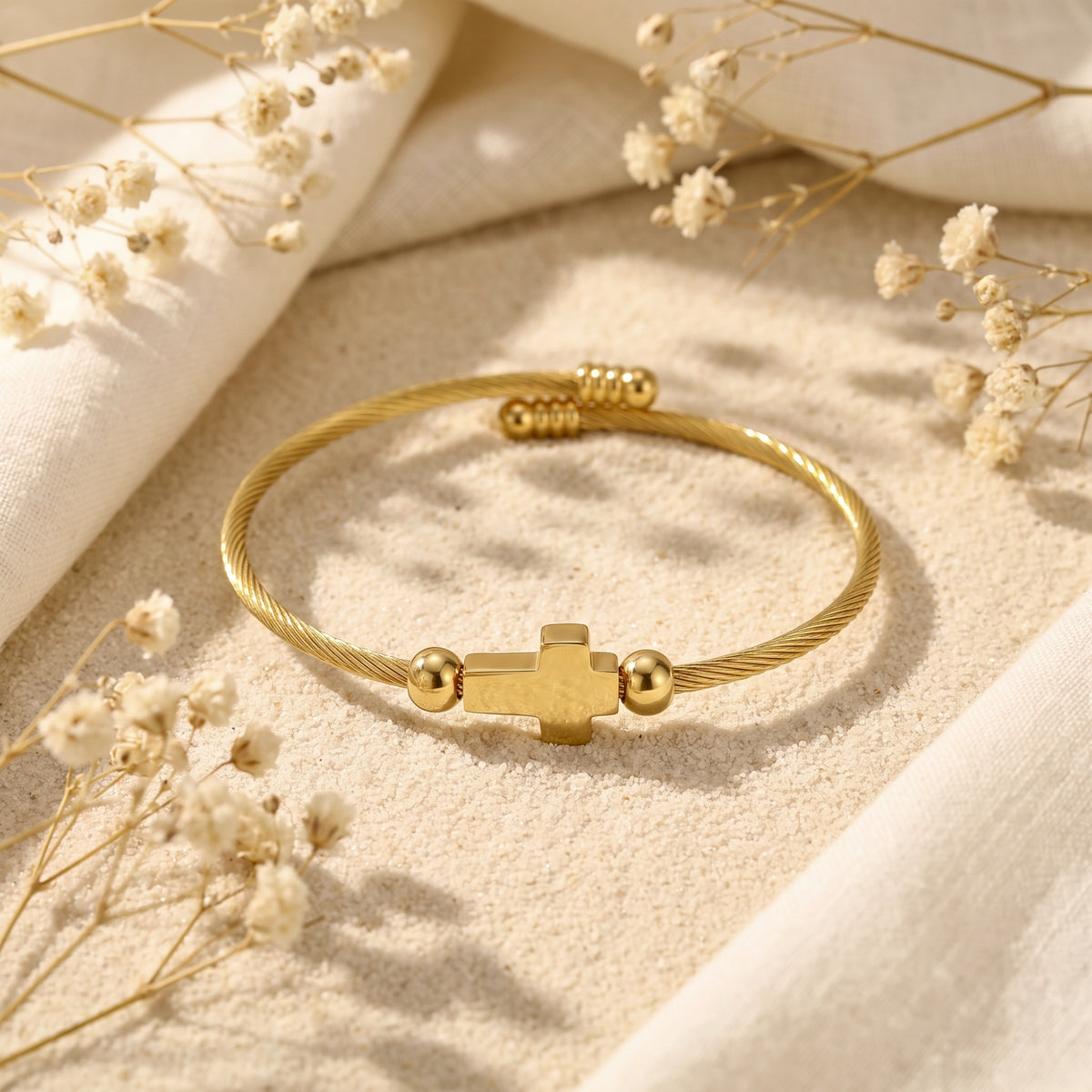 Gold-tone twisted cable cuff bracelet with a polished cross emblem resting on sand beside dried flowers.