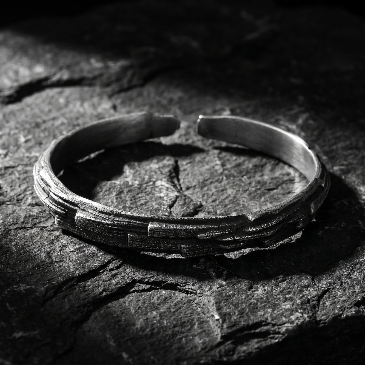 Silver open cuff bracelet with rough, chiseled geological texture displayed on dark slate stone.