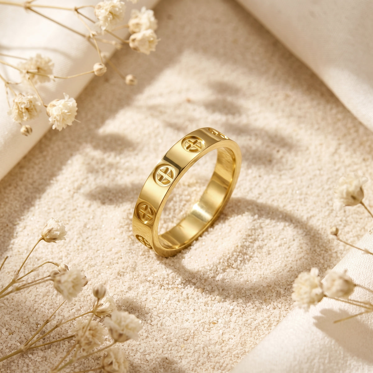 Angled close-up of a gold-tone cross band ring with recessed circular cross motifs standing on fine sand.