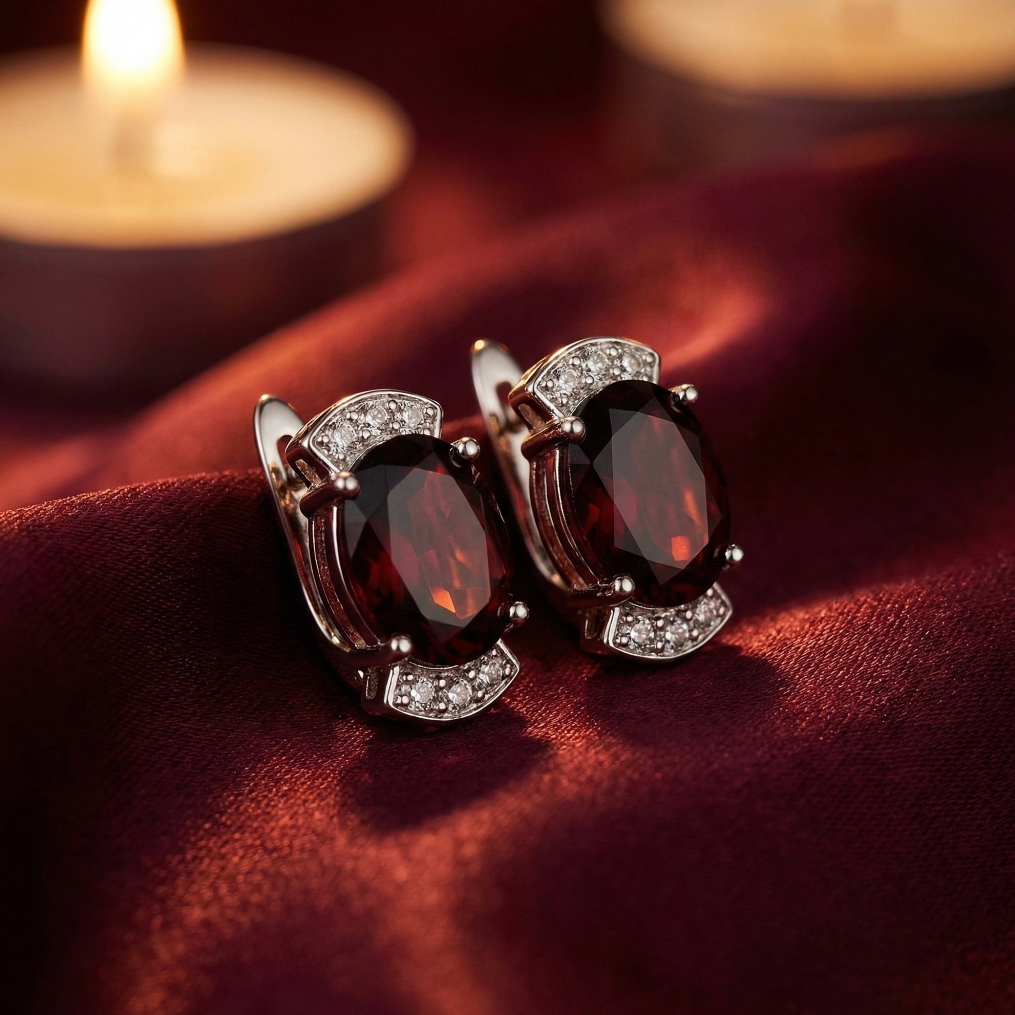 Oval natural garnet drop earrings in sterling silver resting on red silk fabric with a candle background.