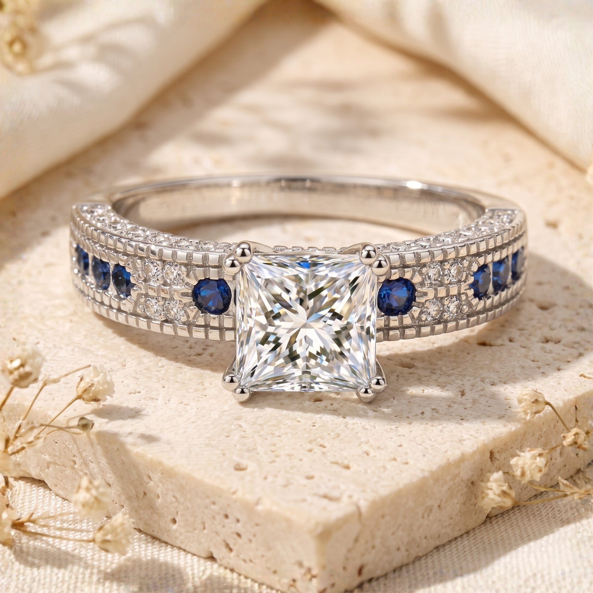 Princess-cut CZ silver ring with blue sapphire accents on a stone surface.