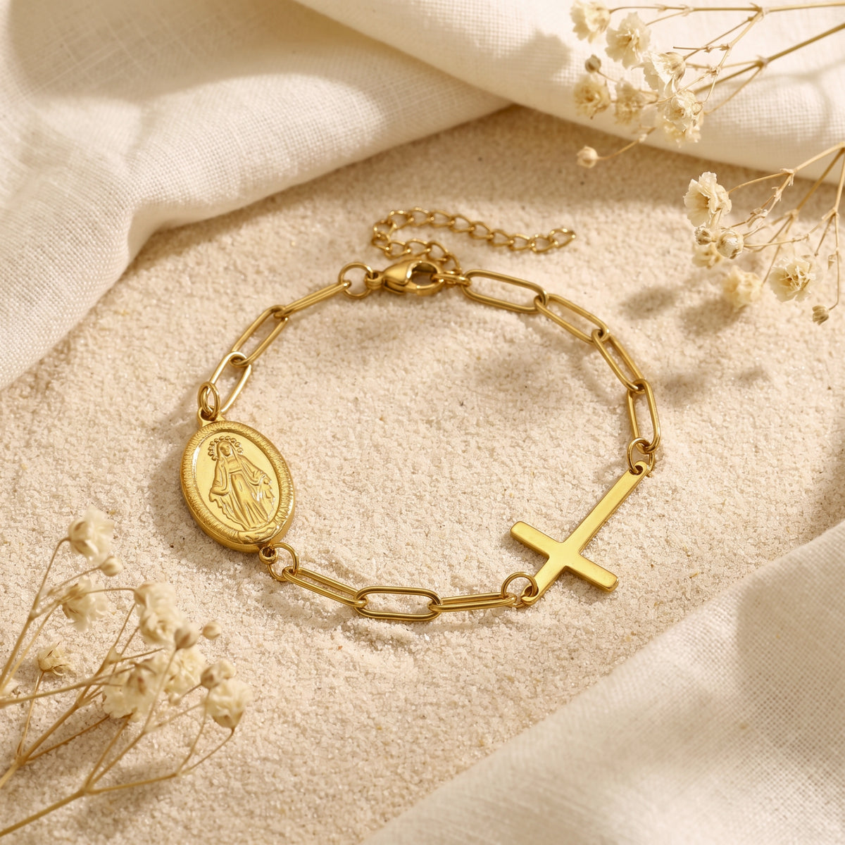 Gold-tone link bracelet with an oval Virgin Mary medal and cross charm on sand, framed by white fabric and dried flowers.