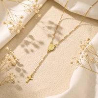 Gold-tone rosary lariat necklace lying on sand with dried white flowers, an oval medal, and a cross drop.