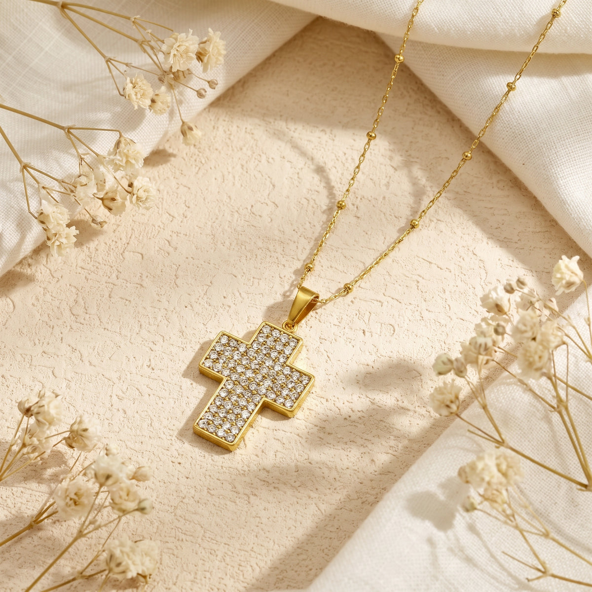 Gold-tone cross pendant with pave stones on a beaded chain, resting on white fabric among dried flowers.