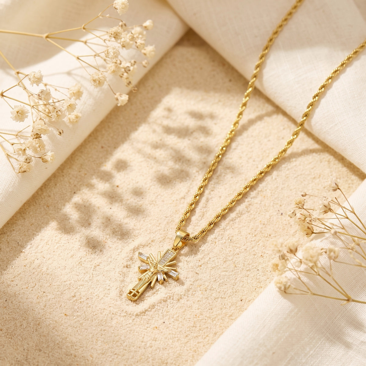 Gold-toned sunburst crucifix pendant and rope chain resting on sand with white fabric and dried flowers.