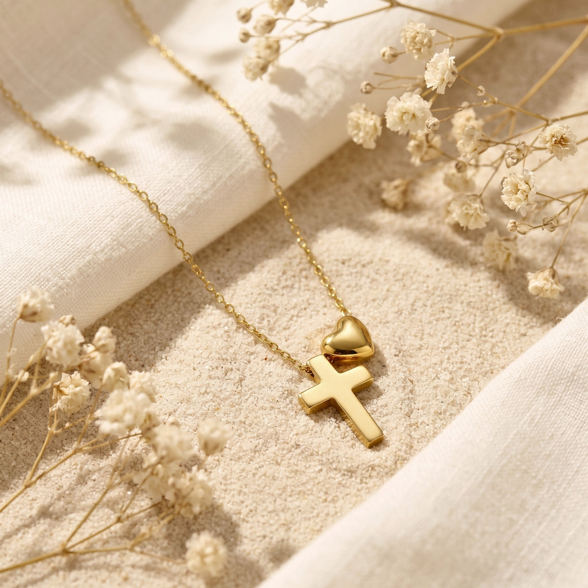 Gold-tone cross and puffed heart necklace resting on sand beside white fabric and dried flowers.